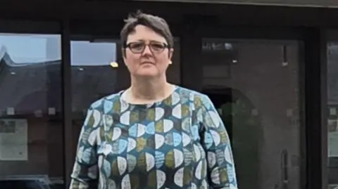 3f9a0ca0-2c33-11f0-822d-6bb6cb319bb5 3f9a0ca0-2c33-11f0-822d-6bb6cb319bb5 A woman with short dark hair and dark glasses, stood in front of a glass building. She is looking into camera and wearing a green, blue and white top. Doctor accused of falsifying artificial mesh records 3f9a0ca0 2c33 11f0 822d 6bb6cb319bb5