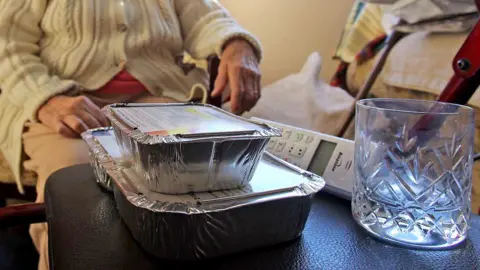 56d09870-2f22-11f0-8ff1-59f5dcf8e9f5 56d09870-2f22-11f0-8ff1-59f5dcf8e9f5 Getty Images Two foil food containers stacked, and empty glass and a landline wireless phone sit on a black leather-like surface. Behind it is an elderly person wearing a beige cable knit cardigan with buttons. Face is not visible and clothing does not make it possible to distinguish if they are male or female. Care providers say overseas worker crackdown 'short-sighted' 56d09870 2f22 11f0 8ff1 59f5dcf8e9f5