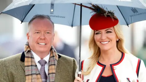 aee8a910-f2cf-11ef-ac99-91c4861fe93d aee8a910-f2cf-11ef-ac99-91c4861fe93d Getty Images A photograph of Doug Barrowman and Baroness Michelle Mone at the Cheltenham horse racing festival in 2019. She is carrying an umbrella and wearing a red hat and white suit. He is wearing a light grey coat and tweed scarf. michelle mone-linked ppe firm evidence to be heard in private Michelle Mone-linked PPE firm evidence to be heard in private aee8a910 f2cf 11ef ac99 91c4861fe93d