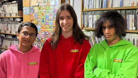 00203af0-4140-11f0-8a46-a5d782130fea 00203af0-4140-11f0-8a46-a5d782130fea Three people in a vintage gaming shop with Pokemon cards and physical video games on shelves behind them. Tushar is on the left with long hair in a ponytail. He’s wearing glasses and smiling, wearing a bright pink “Mario Kart Society” hoodie. Edward is smiling too. He has long hair and wears a red hoodie. Hasan is in a green hoodie and has long ruffled hair. He has a serious look with his arms folded. Stores open at midnight as fans rush to buy Nintendo Switch 2 00203af0 4140 11f0 8a46 a5d782130fea