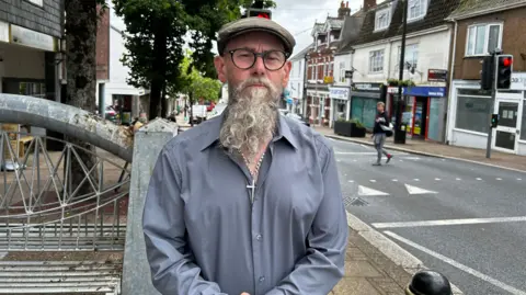 37459db0-4607-11f0-9b27-cd0bc54f64dd 37459db0-4607-11f0-9b27-cd0bc54f64dd A man with a long grey beard, wearing a blue shirt and a brown cap and glasses. He is looking directly at the camera. Behind him to the right is a road and traffic lights. To the left of him is the high street and a metal bench. NHS and education among local spending priorities 37459db0 4607 11f0 9b27 cd0bc54f64dd