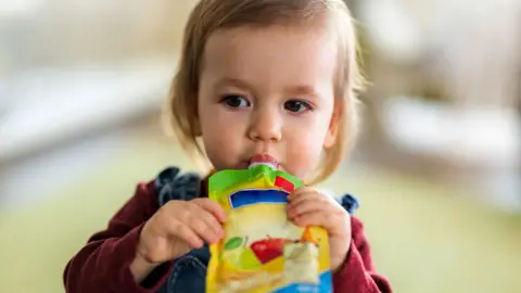 7985b210-20e5-11f0-8c2e-77498b1ce297 7985b210-20e5-11f0-8c2e-77498b1ce297 Getty Images A toddler with blondey-brown hair sucking a pouch of baby food, which she is holding with both hands. She is wearing a burgundy top and dungarees. Restrict shop-bought baby food, government tells parents 7985b210 20e5 11f0 8c2e 77498b1ce297