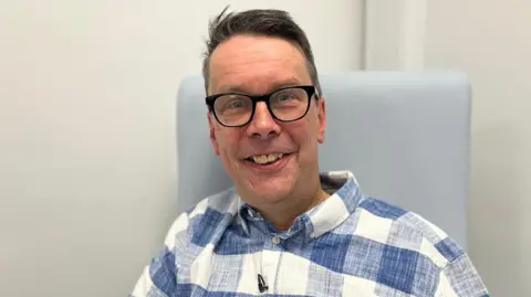 88d18c50-46b3-11f0-98c9-f388968ae068 88d18c50-46b3-11f0-98c9-f388968ae068 BBC Paul Silvester smiling in a chair wearing a blue and white checked linen shirt and black thick-rimmed glasses World-first blood cancer therapy to be given on NHS 88d18c50 46b3 11f0 98c9 f388968ae068