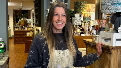 8a4fd5e0-4619-11f0-9942-8ded1e77a695 8a4fd5e0-4619-11f0-9942-8ded1e77a695 A woman with chest length brown hair. She is wearing an apron that is covered in paint and a blue sweater that has paint of the sleeves. Around here is itends from her shop. To the right a wooden chest of drawers with a lamp and to the right a grey chest of drawers with notebooks on. NHS and education among local spending priorities 8a4fd5e0 4619 11f0 9942 8ded1e77a695