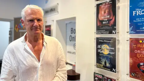 96dcfaf0-4c4e-11f0-ae71-69df9e8fb45a 96dcfaf0-4c4e-11f0-ae71-69df9e8fb45a Tom Binet has gray hair, wearing a white shirt, stands in front of a wall displaying various event posters. The posters include titles like 'Below the Belt,' 'Frozen,' and 'Music at the Orangerie.' Above the posters is a sign that reads 'this week.' In the background, there is a box office sign and another poster partially visible through an open doorway. Calls for more support for endometriosis patients 96dcfaf0 4c4e 11f0 ae71 69df9e8fb45a