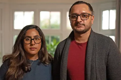 aacff360-cf6b-11ef-87df-d575b9a434a4 aacff360-cf6b-11ef-87df-d575b9a434a4 MARTIN MCQUADE / BBC Amarjit and Mandip pictured standing next to each other. Amarjit has long brunette hair and black-rimmed glasses. She is wearing a blue jumper and a silver necklace. Mandeep has dark hair which is tied back, black-rimmed glasses and a short beard. He is wearing a red t-shirt and grey woollen cardigan. They are pictured in front of a white-framed window with green plants outside. Leeds maternity units downgraded to 'inadequate' aacff360 cf6b 11ef 87df d575b9a434a4