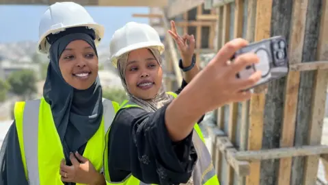 aecaea30-46c9-11f0-bbaa-4bc03e0665b7 aecaea30-46c9-11f0-bbaa-4bc03e0665b7 Anthony Irungu / BBC Saadia Ahmed Omar (right) takes a photo of herself and Fathi Mohamed Abdi (left) atop a building under construction in Mogadishu. They are both wearing hard hats over their headscarves and are in high vis yellow vests. Ms Omar makes the victory sign as she takes the photo. The women at the centre of Somalia's construction boom aecaea30 46c9 11f0 bbaa 4bc03e0665b7
