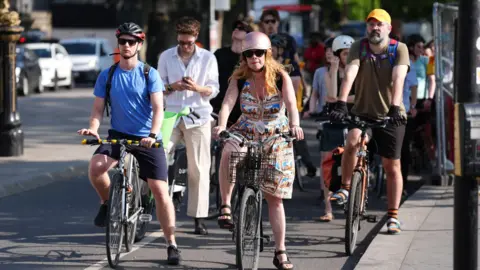 cfc3aac0-4cf9-11f0-b48b-8b6357a99d04 cfc3aac0-4cf9-11f0-b48b-8b6357a99d04 PA Media Cyclists pause on the road, some wearing helmets, as the sun shines down. Amber heat health alerts in place as temperatures above 30C likely cfc3aac0 4cf9 11f0 b48b 8b6357a99d04