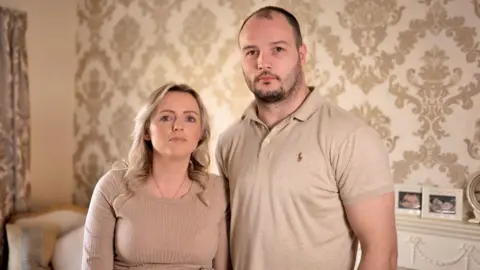f2929db0-45fe-11f0-bace-e1270fc31f5e f2929db0-45fe-11f0-bace-e1270fc31f5e Heidi and Dale standing close together in a living room with cream patterned wallpaper behind them. Pregnancy scan photos are framed on the fireplace. Heidi has long, wavy blonde hair and is wearing a ribbed, long sleeved top and gold necklace. Dale has short dark hair with a beard and is wearing a cream polo-shirt. Dozens more parents demand maternity care inquiry f2929db0 45fe 11f0 bace e1270fc31f5e