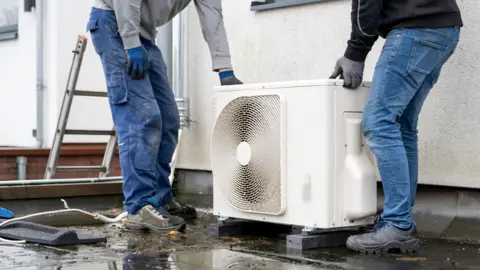 fcc73d70-3bc4-11f0-bb14-a341f4f72a30 fcc73d70-3bc4-11f0-bb14-a341f4f72a30 Getty Images The shot shows a white heat pump being installed on a wet flat roof by two men in jeans and boots. Just the bottom half of their bodies are visible as they fit the pump into place. A ladder is visible just to the left of the picture. Planning change to make installing heat pump easier for millions fcc73d70 3bc4 11f0 bb14 a341f4f72a30