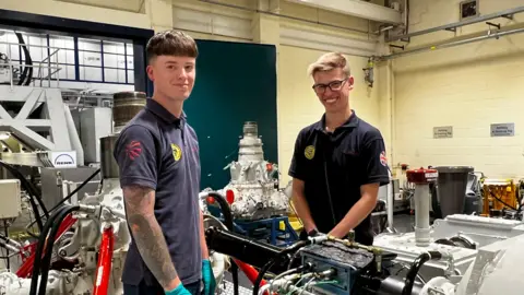 0cf3c9b0-6312-11f0-a700-fb7b15f3374f 0cf3c9b0-6312-11f0-a700-fb7b15f3374f BBC Two men in a factory smile at camera. There are various engine parts behind them. Both are wearing blue tops, and are younger workers. The factory walls are painted yellow. Defence firms 'confident' hundreds more jobs will come 0cf3c9b0 6312 11f0 a700 fb7b15f3374f
