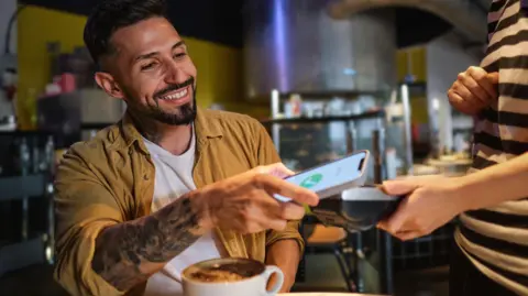 3c2d0f80-581c-11f0-8b01-cfb61de133b2 3c2d0f80-581c-11f0-8b01-cfb61de133b2 Getty Images Smiling young man in a cafe pays using a phone with a coffee in front of him. British Bulldogs to mobile phones – your banknote redesign ideas 3c2d0f80 581c 11f0 8b01 cfb61de133b2