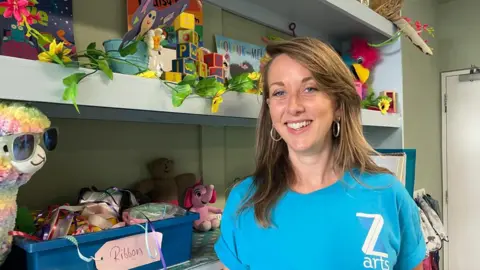 Elaine Doran/BBC Gilly Balfour with shoulder length brown hair wearing a blue t shirt with the logo Z arts on it smiling in front of shelves of colourful children's games and art materials  Summer holiday club costs: &#039;It&#039;s the amount you&#039;d spend going away&#039; 3cdb3e80 625c 11f0 82c0 a54b0ccf49e8