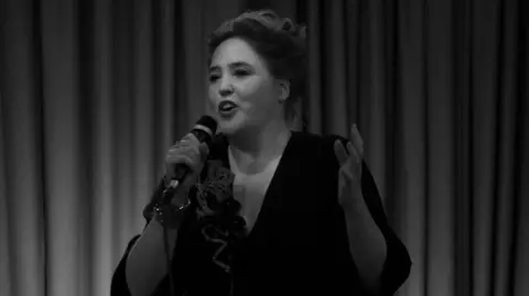 Scary Little Girls A black and white photo. A woman wearing a black dress. She is holding a microphone in her hand. She has her hair up in a bun. There are curtains behind her.   Cornwall theatre company awarded nearly £20,000 4635bd80 5be9 11f0 9584 0dcfb55e8671