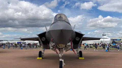 466afc20-632d-11f0-8a13-91ee44fd4116 466afc20-632d-11f0-8a13-91ee44fd4116 A military fighter jet, stands on a red runway. Behind it, behind fencing, crowds of people walk past and look on. There are tents behind them. Defence firms 'confident' hundreds more jobs will come 466afc20 632d 11f0 8a13 91ee44fd4116