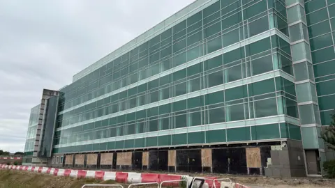 Ben Schofield/BBC Modern multi-story building with glass windows, the ground floor us under construction with boarded-up sections.  New science centre aims to help fight pandemics 51f3ca50 62f0 11f0 83d2 4f671b8c1523