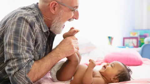 Getty Images An older dad with a grey beard and wearing glasses and a checked shirt, holds a young baby's feet while she lies on a bed, wearing a nappy, and smiles up at him  &#039;Notable&#039; rise in dads over 60 in England and Wales c0ba57e0 5677 11f0 95fc edf89039c20a