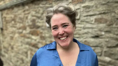 BBC A woman looking at the camera and smiling. She has short hair. She is wearing a blue top. There is a blurred brick wall behind her.  Cornwall theatre company awarded nearly £20,000 d791a680 5bf4 11f0 9584 0dcfb55e8671