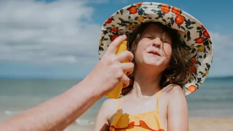 e7e2c360-4b6a-11f0-abc3-41f059ca87b9 e7e2c360-4b6a-11f0-abc3-41f059ca87b9 Getty Images A little girl on the beach wearing a sun hat and orange swimming costume grimaces as sunscreen is applied to her face. What should you look for when choosing sunscreen? e7e2c360 4b6a 11f0 abc3 41f059ca87b9